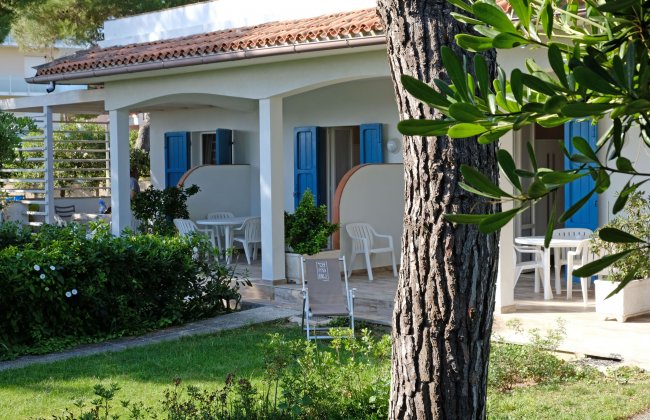 Bungalow bianco con finestre blu, veranda e sedie su un prato verde.