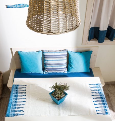 Tavolino con centrino bianco e blu, cuscini coordinati su una panca, lampadario di vimini.
