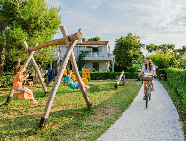 Bambini su altalene in un giardino, persona in bici su vialetto di ghiaia.