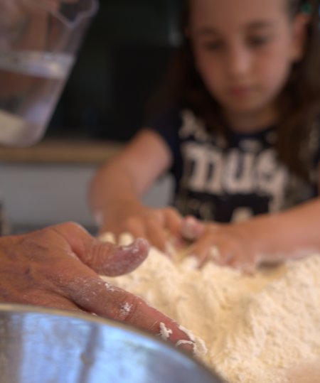 Mani che impastano farina, con una bambina sullo sfondo che osserva.