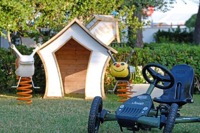Un kart giocattolo su prato, vicino a giochi a forma di lumaca e ape e una casetta di legno.