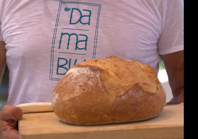 Persona con maglietta bianca e scritta, tenendo un pane casereccio su un tagliere.