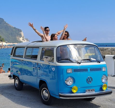 Furgoncino Volkswagen blu vicino al mare. Due persone sporgono dal tetto, godendosi la giornata.