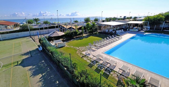 Vista di una piscina esterna con lettini e un campo da tennis vicino al mare.