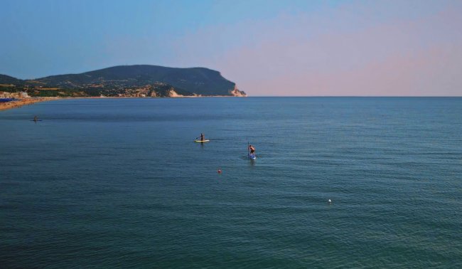 Mare calmo con persone che praticano surf, e una montagna sullo sfondo.