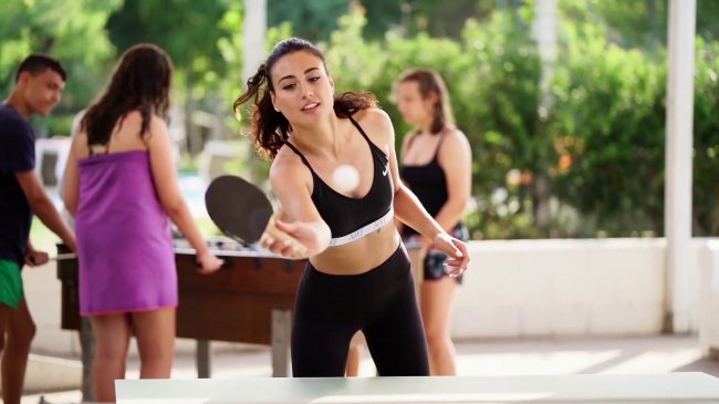 Donna gioca a ping pong all'aperto con altre persone sullo sfondo.