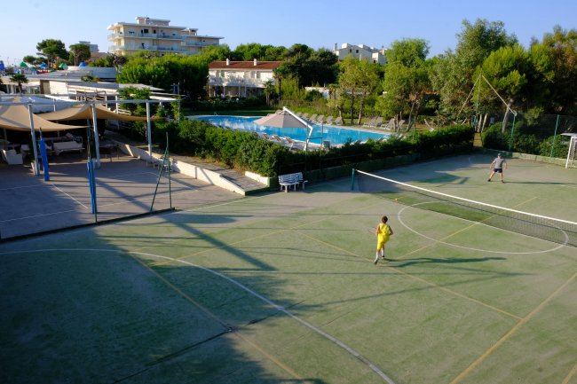 Campo da tennis all'aperto con persone che giocano; piscina e giardini vicini.