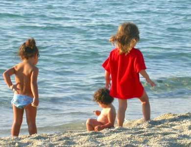 Tre bambini giocano sulla spiaggia vicino all'acqua; uno indossa una maglietta rossa.