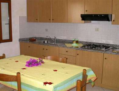 Cucina con tavolo coperto da tovaglia gialla, armadietti in legno chiaro e fornelli.