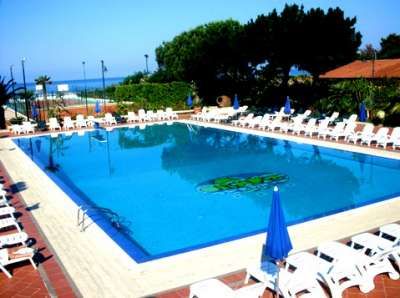 Una grande piscina rettangolare con sdraio e ombrelloni blu intorno, vista sul mare.