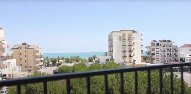 Vista su edifici alti, alberi e mare in lontananza, osservata da un balcone.