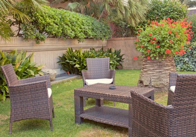 Giardino con sedie e tavolino in vimini, circondato da piante e un cespuglio fiorito.