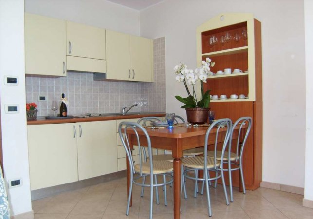 Cucina con mobili chiari, tavolo in legno e sedie, fiore al centro.