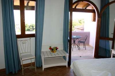 Camera con tende blu, finestra e porta aperta su un balcone con tavolo e sedie.
