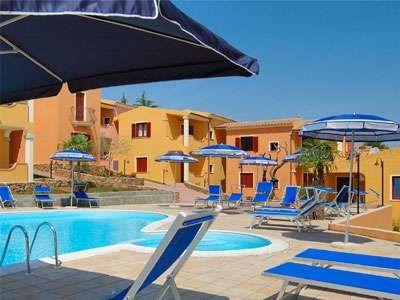 Piscina circondata da lettini e ombrelloni blu, edifici colorati sullo sfondo.