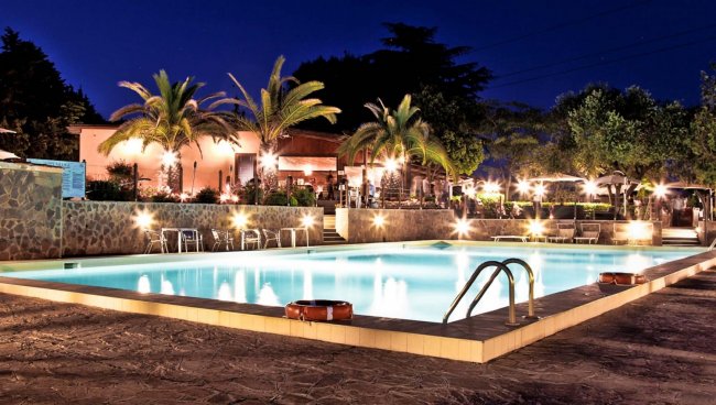 Piscina illuminata di sera con palme e luci decorative.