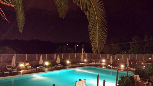 Piscina illuminata di notte con ombrelloni e palme.