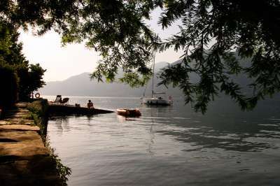 Un molo sull'acqua con persone sedute sotto un albero. In lontananza, una barca a vela sul lago.