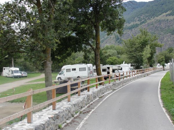 Una strada con camper parcheggiati accanto a un prato verde e alberi, con montagne sullo sfondo.