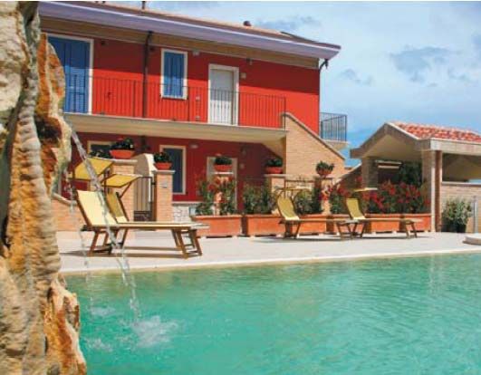 Piscina con cascata, lettini e edificio rosso sullo sfondo.