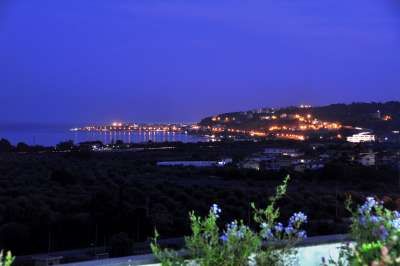 Vista notturna di una costa illuminata con colline sullo sfondo e luci lontane.