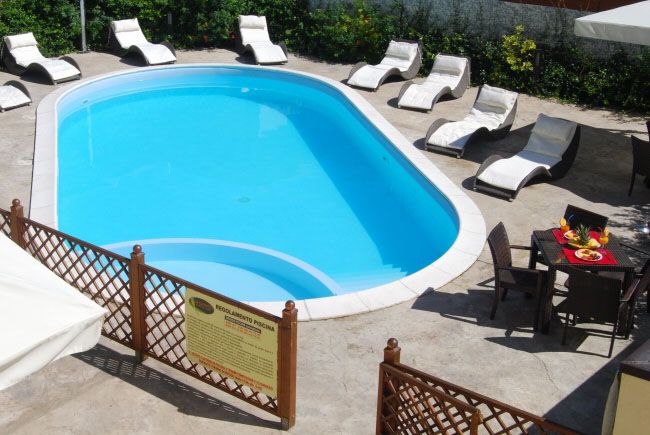 Piscina ovale circondata da lettini bianchi, con tavolo e sedie a lato.