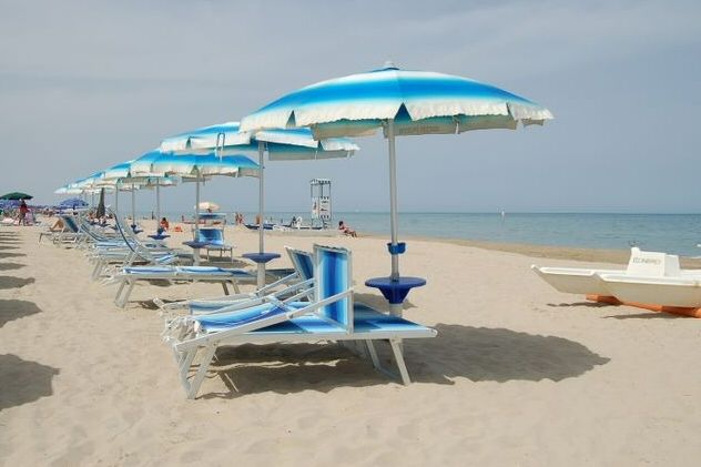 Spiaggia con ombrelloni e lettini blu su sabbia chiara, mare calmo sullo sfondo.