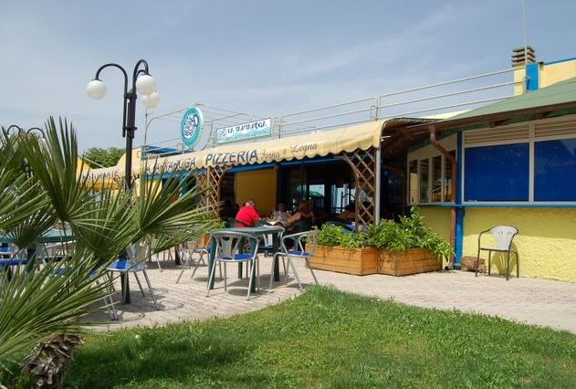 Esterno di una pizzeria con tavoli all'aperto, un'insegna blu e verde, e piante decorative.