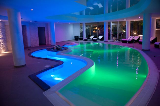 Piscina interna con illuminazione colorata e lettini bianchi.