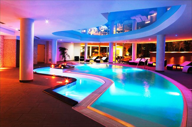 Piscina interna con illuminazione colorata, lettini neri e ampie finestre con piante.