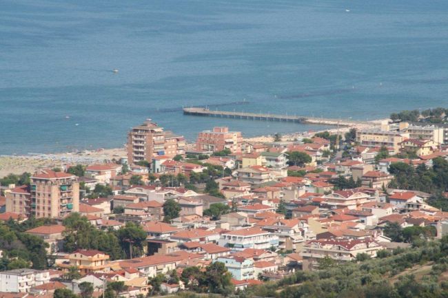 Vista di una città costiera con edifici dai tetti rossi e un molo sul mare.
