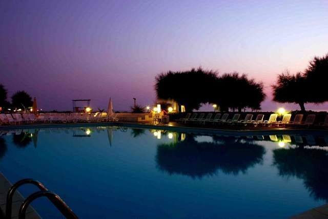 Piscina illuminata al tramonto con sdraio allineate e alberi sullo sfondo.