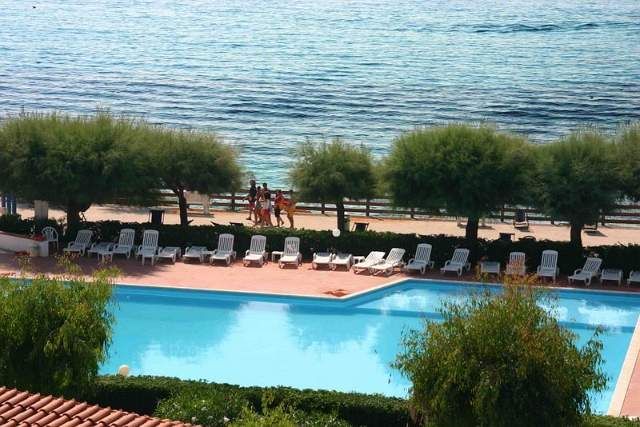 Piscina azzurra vista dall'alto, vicino al mare con sdraio bianche e alberi.