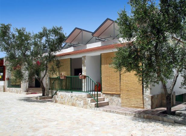 Bungalow bianco con tende di bambù, terrazza, alberi su strada di pietra.