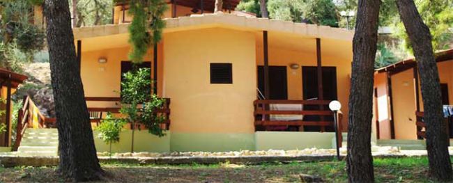 Bungalow giallo con terrazza in legno, circondato da alberi.