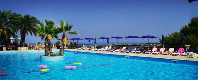 Piscina con palme al centro, sedie a sdraio e ombrelloni blu sullo sfondo.