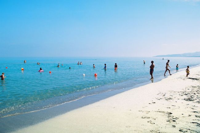Persone che nuotano e camminano lungo una spiaggia con mare calmo e limpido sotto un cielo azzurro.