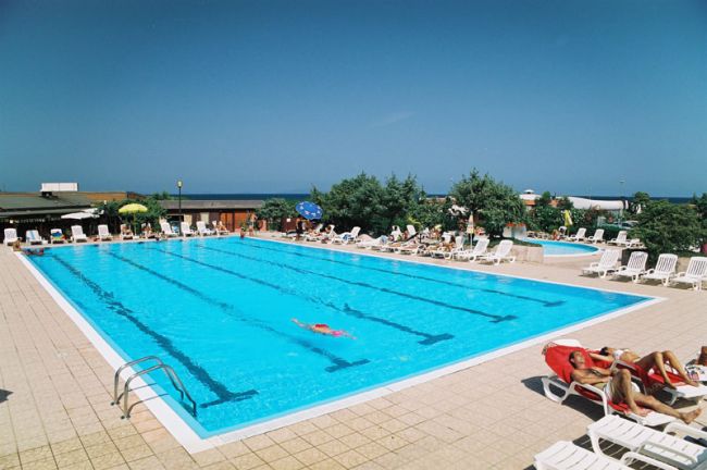 Ampia piscina con corsie, persone che nuotano e rilassano sulle sdraio sotto un cielo blu.