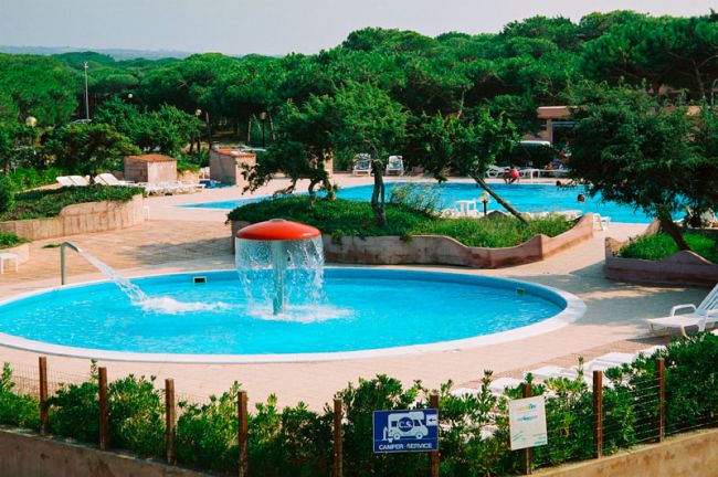 Una piscina circolare con giochi d'acqua, circondata da verde e lettini.
