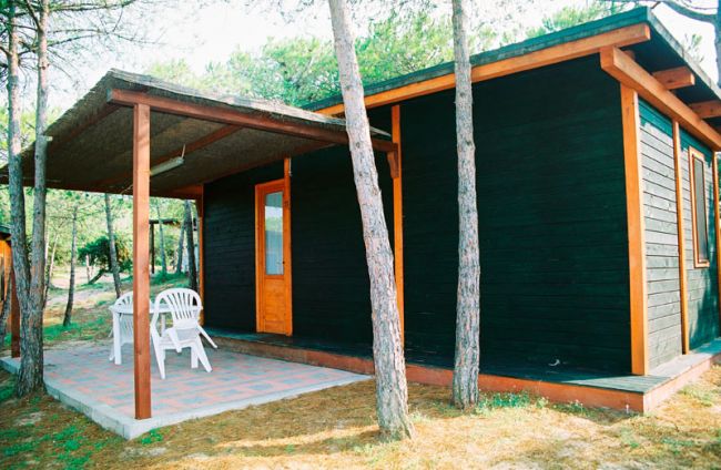 Casetta in legno scuro con veranda, tavolo e sedie bianche all'esterno, circondata da alberi.