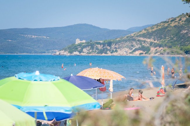 Spiaggia con ombrelloni colorati e bagnanti, mare azzurro e colline sullo sfondo.