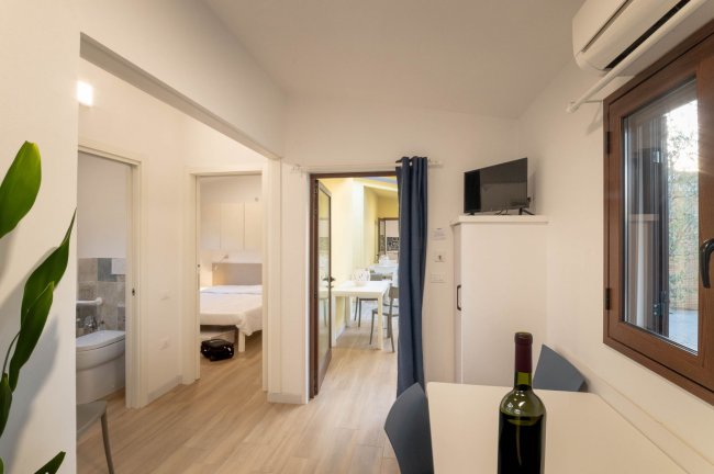 Un interno moderno con sala, tavolo, bottiglia di vino, camera e bagno visibili con parquet chiaro.