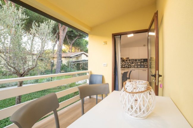 Balcone soleggiato con tavolo bianco e sedie, vista su giardino alberato, porta aperta su cucina.