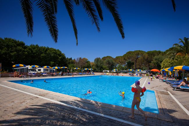Bambini giocano in piscina con ombrelloni colorati intorno.