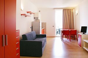 Salotto luminoso con tavolo rosso, divano blu e accesso a un balcone.