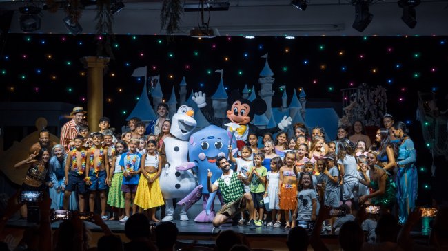 Gruppo di bambini e mascotte su un palco colorato.