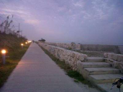 Una strada pedonale al tramonto con lampioni accesi e un muretto di pietra sulla destra.