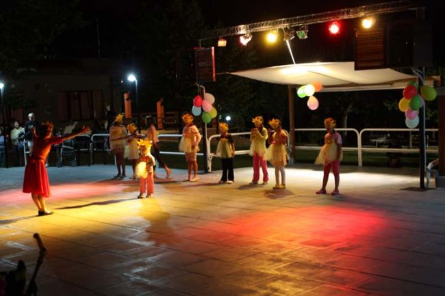 Bambini in costume seguono una donna in rosso su un palco illuminato.