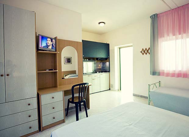 Camera da letto con mobili semplici e TV accesa.
