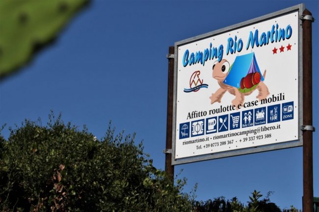 Cartello del Camping Rio Martino tra la vegetazione.